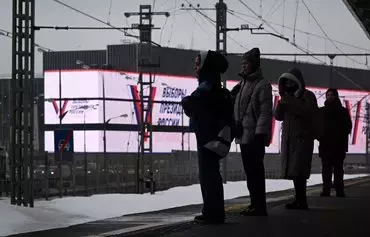 Passengers wait for a train at a station of the Moscow Central Ring, a commuter rail line circling the city, with a huge advertising screen announcing the 2024 presidential election in the distance, in Moscow on December 18. [Natalia Kolesnikova/AFP]