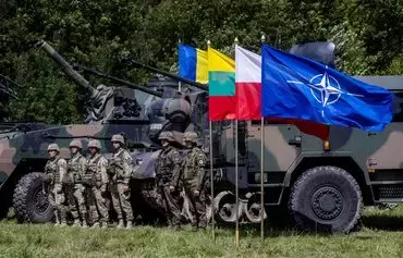 Польские (справа) и румынские (слева) солдаты рядом с военными машинами и флагом НАТО на полях пресс-конференции польского и литовского президентов после посещения лидерами мобильного командного центра Многонациональной дивизии НАТО «Северо-Восток», расположенного в так называемом Сувалкском коридоре — зажатой между российской Калининградской областью и Беларусью 80-километровой полосе польско-литовской границы. Поселок Шиплишки, северо-восток Польши, 7 июля 2022 г. [Войтек Радвански/AFP]