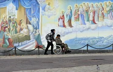 A girl carries a wheelchair with a disabled serviceman in front of frescoes of St. Michael's Golden-Domed Cathedral in Kyiv on November 20, 2023, amid the Russian invasion of Ukraine. [Sergei Supinsky/AFP]